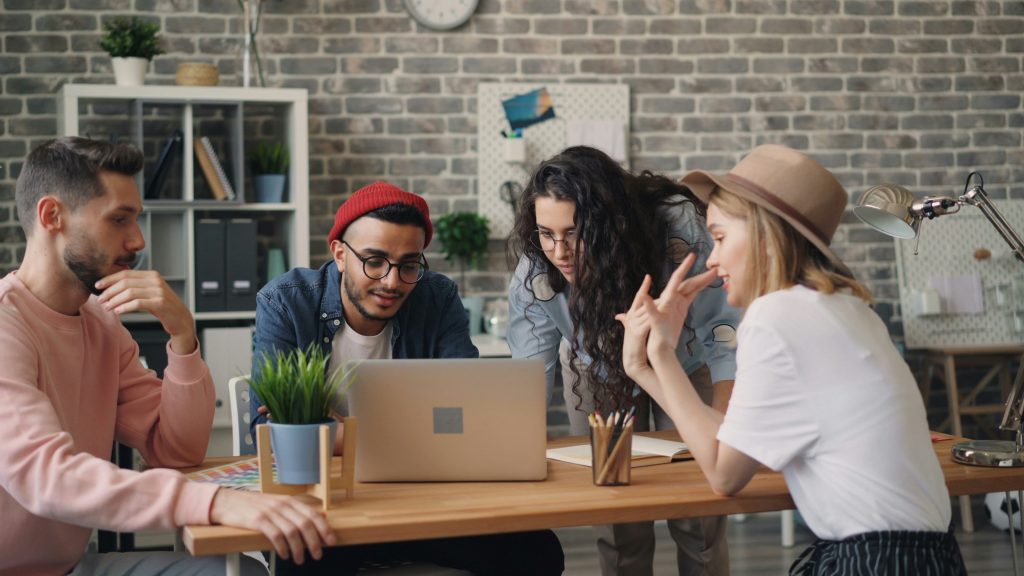 Eine Gruppe von vier jungen, kreativen Fachleuten arbeitet gemeinsam an einem Holztisch in einem modernen Büro mit Ziegelwand. Zwei Männer und zwei Frauen sind in eine Diskussion vertieft, wobei ein Mann auf einen Laptop blickt und eine Frau lebhaft gestikuliert. Die Szene vermittelt Kollaboration und das gemeinsame Gestalten von Prozessen und Workflows.