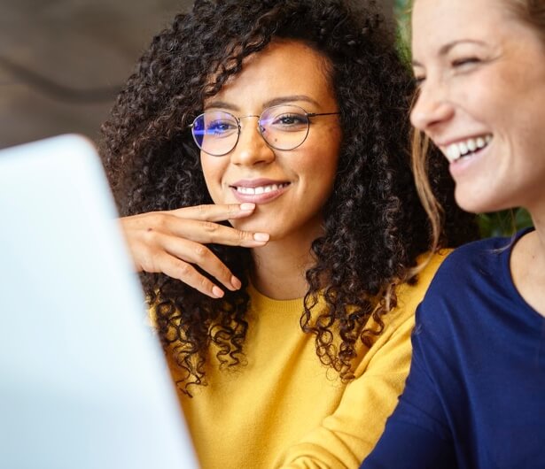 Zwei lachende Frauen, die zusammen auf einen Laptop schauen; die Frau im gelben Pullover trägt eine Brille und hält eine Hand am Kinn.