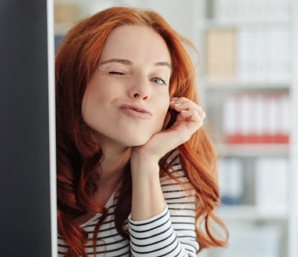 Fröhliche Frau mit langen roten Haaren, die schelmisch zwinkert und den Kopf auf die Hand stützt, während sie aus dem Bildrand blickt.