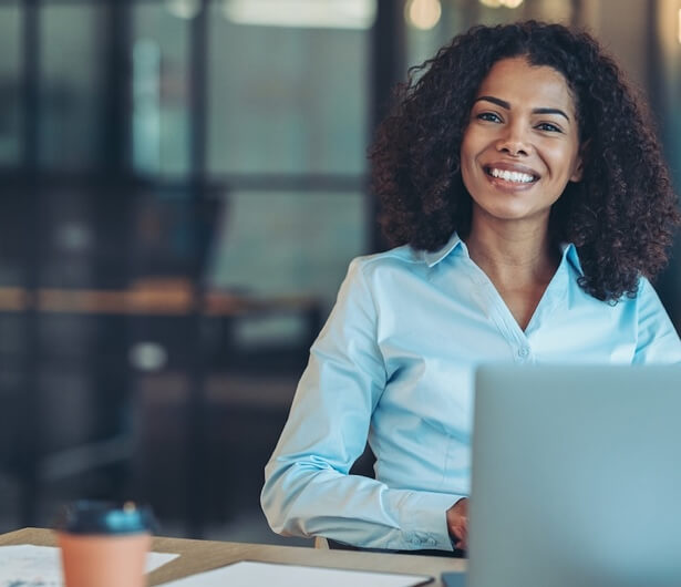 Lächelnde junge Geschäftsfrau mit dunklen Locken, die am Schreibtisch vor einem Laptop sitzt.